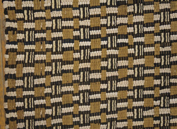 Low-profile textured rug with a black, beige, and tan geometric woven pattern on a wooden floor.