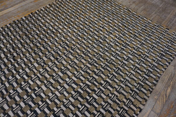 A low-pile woven rug featuring a black, beige, and off-white checkered pattern rests on gray wood flooring.
