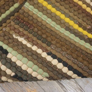 Chunky braided rug with diagonal stripes in earth tones, green, yellow, and cream, resting on distressed wood.