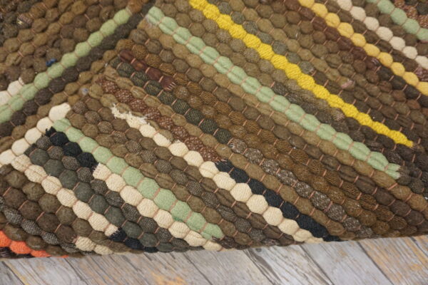 Chunky braided rug with diagonal stripes in earth tones, green, yellow, and cream, resting on distressed wood.
