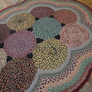 Braided oval rug displaying colorful speckled circles and rings, lying on rustic wooden floorboards.
