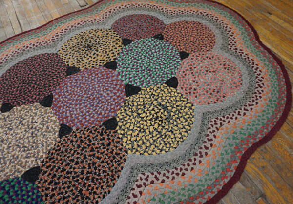 Braided oval rug displaying colorful speckled circles and rings, lying on rustic wooden floorboards.