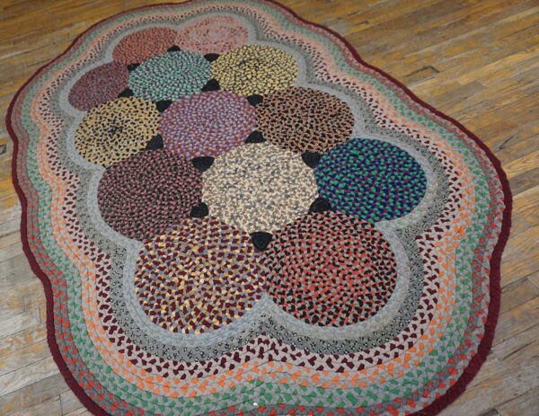 Multicolored oval braided rug with large speckled circles and banded borders resting on light wood flooring.