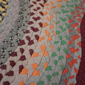Braided rug detail featuring concentric rows of multicolor fabric patches on distressed gray and yellow wood flooring.