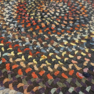 Close-up texture of a multicolored braided rug featuring concentric bands of dark gray, orange, and purple.