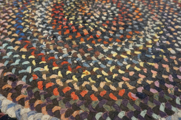 Close-up texture of a multicolored braided rug featuring concentric bands of dark gray, orange, and purple.