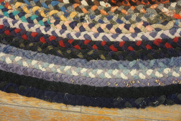 Thick multicolored braided rug featuring concentric bands of blue, black, red, and neutral tones on wood flooring.