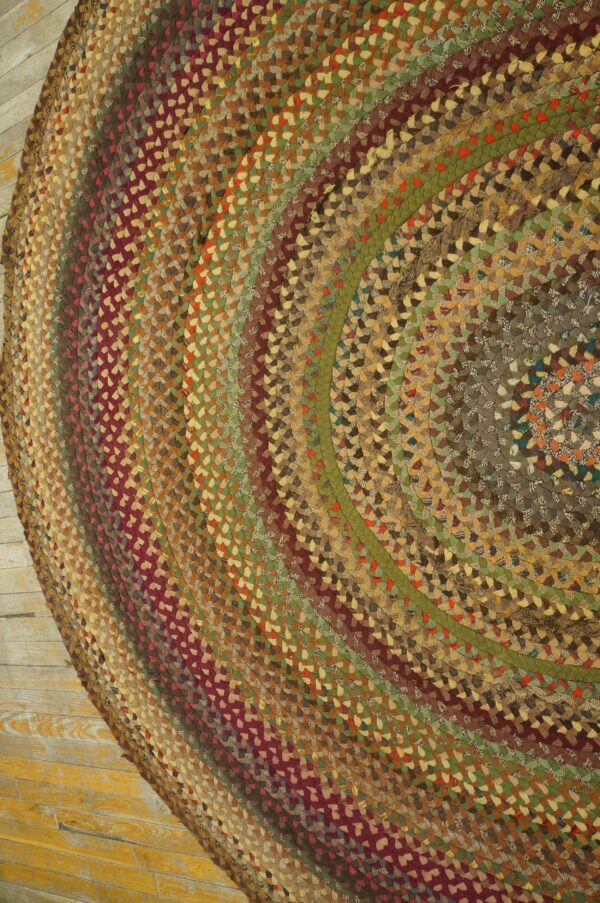 Braided rug featuring concentric bands of mixed maroon, brown, green, and tan fabric on wood flooring.