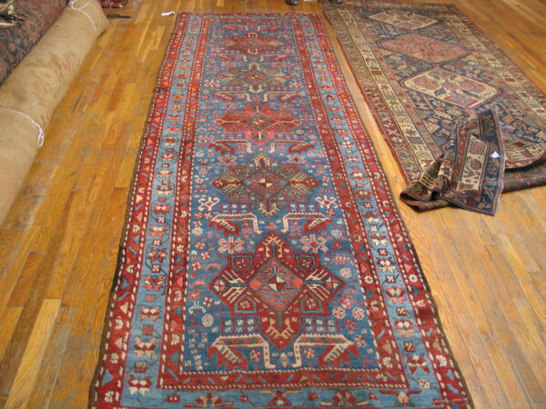 Geometric blue and red patterned runner rug laid flat on hardwood flooring alongside other stored rugs.