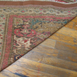 Geometric brown, pink, and beige flatwoven rug partially covers worn, yellowish wood plank flooring.