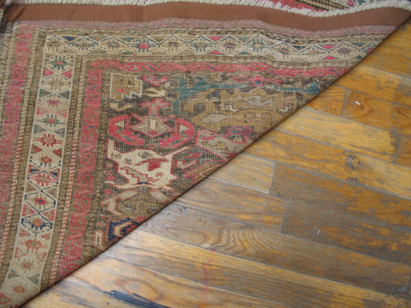 Geometric brown, pink, and beige flatwoven rug partially covers worn, yellowish wood plank flooring.