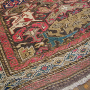 Detailed corner of a low-pile rug featuring complex geometric patterns in brown, red, tan, and teal on a wood floor.
