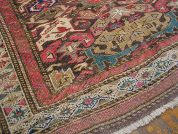 Detailed corner of a low-pile rug featuring complex geometric patterns in brown, red, tan, and teal on a wood floor.
