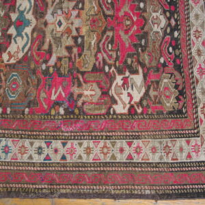 Intricately patterned rug corner featuring geometric designs and stylized figures in magenta, brown, and cream on wood flooring.