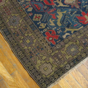 Corner view of a low-pile blue rug with red and olive geometric patterns resting on light hardwood planks.
