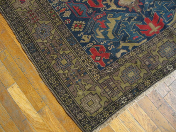 Corner view of a low-pile blue rug with red and olive geometric patterns resting on light hardwood planks.