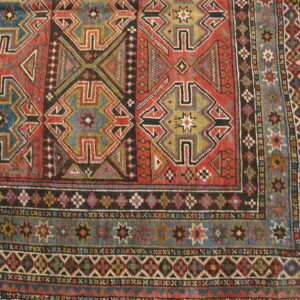 Geometric rug corner detail showing stepped designs in red, teal, black, and gold on a plank wood floor.