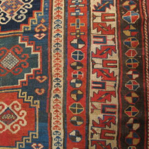 Tightly woven rug with bold geometric patterns in red, navy, teal, and cream on a wooden floor.