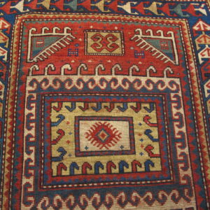 Geometric rug in red, blue, and tan with intricate borders and a stepped central medallion on wood planks.