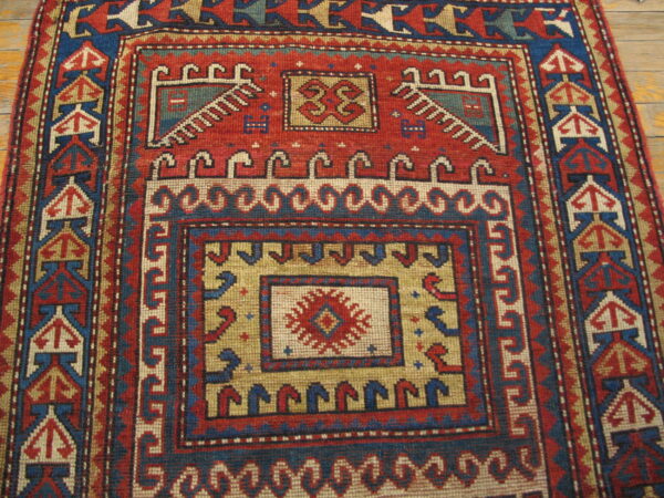 Geometric rug in red, blue, and tan with intricate borders and a stepped central medallion on wood planks.