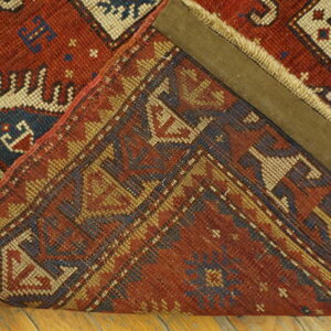 Red and blue geometric rug is folded over, displaying the knotted reverse side on wood flooring.