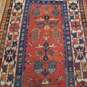 Low-pile tribal rug with rust field, geometric blue borders, and multi-colored medallions on wood flooring.