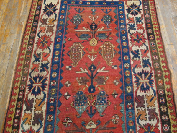 Low-pile tribal rug with rust field, geometric blue borders, and multi-colored medallions on wood flooring.