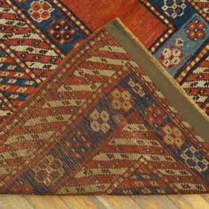 Geometric red, blue, and cream rug with diagonal stripes folded over, resting on light wood floorboards.