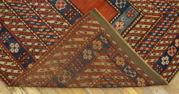 Geometric red, blue, and cream rug with diagonal stripes folded over, resting on light wood floorboards.