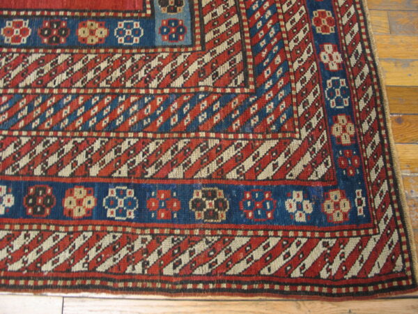 Red and blue geometric rug border with diagonal stripes and stylized medallions on a natural wood floor.