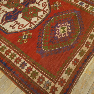 Red flatweave rug with geometric patterns in blue, purple, olive, and cream on distressed wooden floorboards.