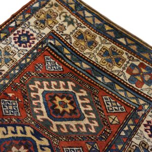 Corner detail of a patterned rug in red, blue, and cream with complex geometric borders.