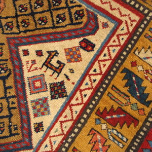 Corner detail of a tribal rug showing mustard, navy, red, and cream geometric patterns and stylized figures.