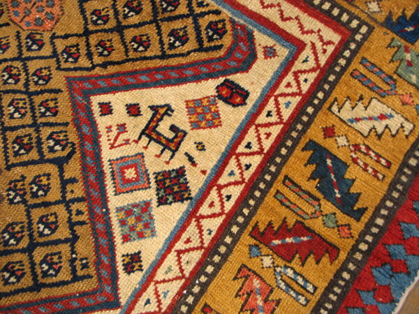 Corner detail of a tribal rug showing mustard, navy, red, and cream geometric patterns and stylized figures.