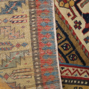 Geometric red, tan, and blue low-pile rug edge curled over, revealing the flat woven reverse side on wood flooring.