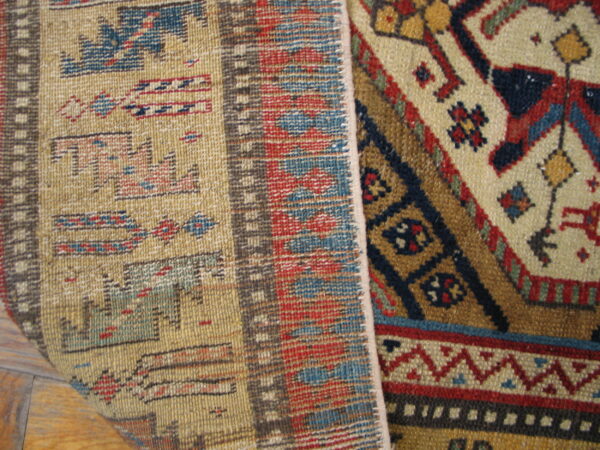 Geometric red, tan, and blue low-pile rug edge curled over, revealing the flat woven reverse side on wood flooring.