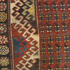 Geometric rug close-up in red, cream, blue, and brown, showing repeating borders next to wood.