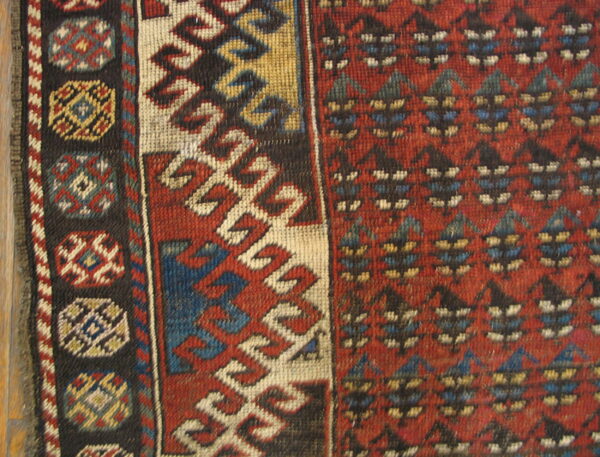 Geometric rug close-up in red, cream, blue, and brown, showing repeating borders next to wood.