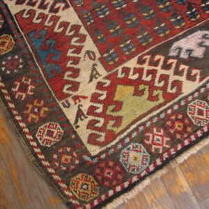 Corner of a low-pile geometric rug in red, dark brown, and cream on rustic wood floorboards.