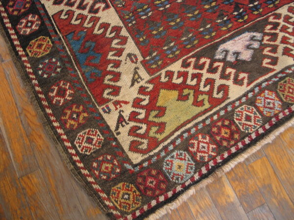 Corner of a low-pile geometric rug in red, dark brown, and cream on rustic wood floorboards.