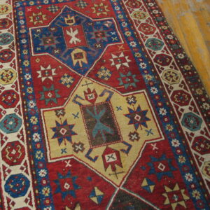 Red and blue tribal rug with geometric medallions and intricate borders rests on worn hardwood flooring.