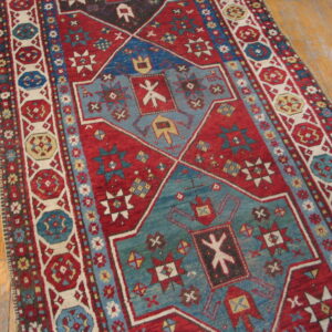 Red and blue geometric pattern rug with cream accents displayed on worn light hardwood floor.