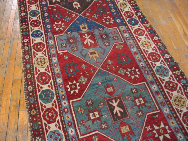 Red and blue geometric pattern rug with cream accents displayed on worn light hardwood floor.