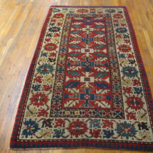 Red field rug with navy blue and cream geometric patterns lays on light brown wood planks.