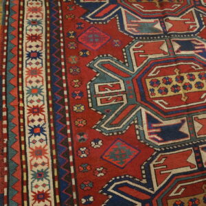 A red rug with complex blue, teal, and cream geometric designs lies on light wood planks.
