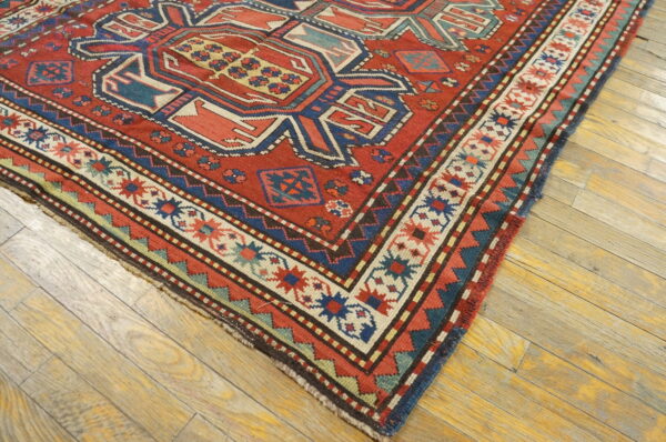 Low-pile red rug with complex blue and cream geometric designs lies on light distressed hardwood planks.