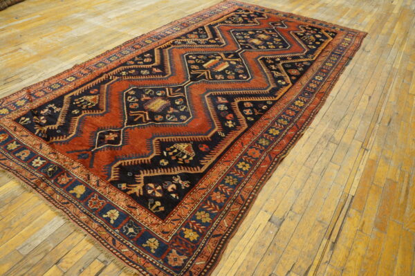 Rust and navy geometric rug featuring stepped diamond patterns rests on worn wood flooring.