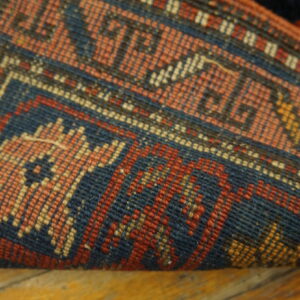A curled rug edge shows tightly woven blue, red, and peach geometric designs over wood flooring.