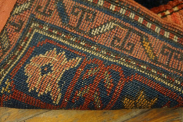 A curled rug edge shows tightly woven blue, red, and peach geometric designs over wood flooring.