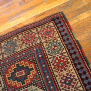 Geometric red, blue, and tan patterned rug lies diagonally on light brown wooden plank flooring.